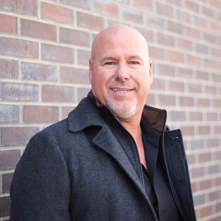 portrait of John Gagne, CEO and founder of Elevation Ten Thousand, wearing a gray coat against a brick background