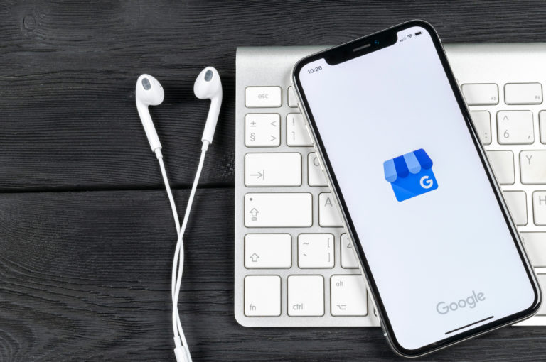 black iPhone with Google My Business app loading placed on a silver Mac keyboard next to white earbuds